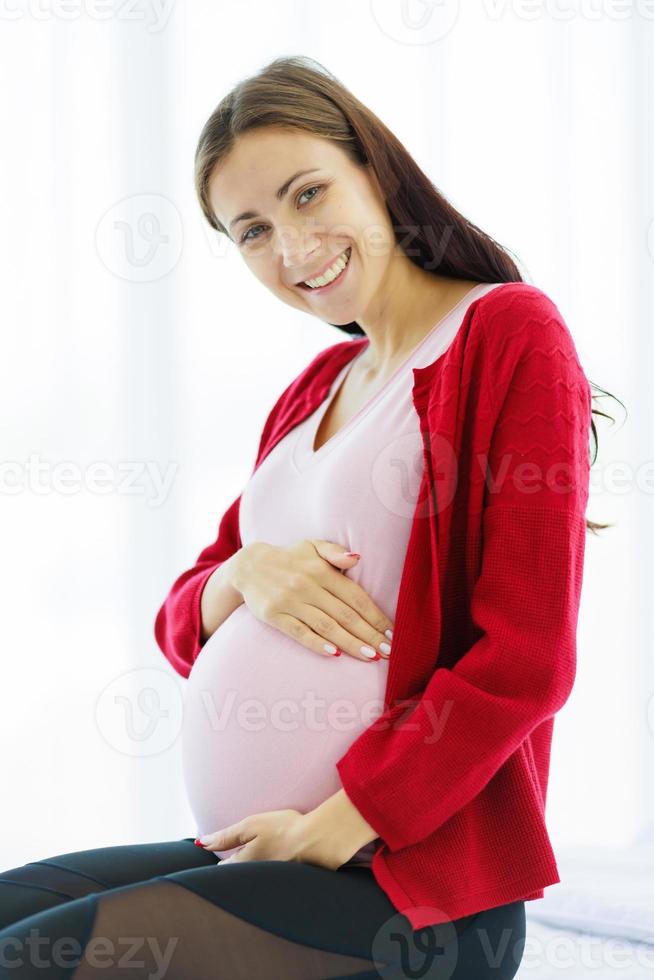 Pregnant woman sits on the bed in her bedroom, caressing her belly with a happy, smiling face. photo