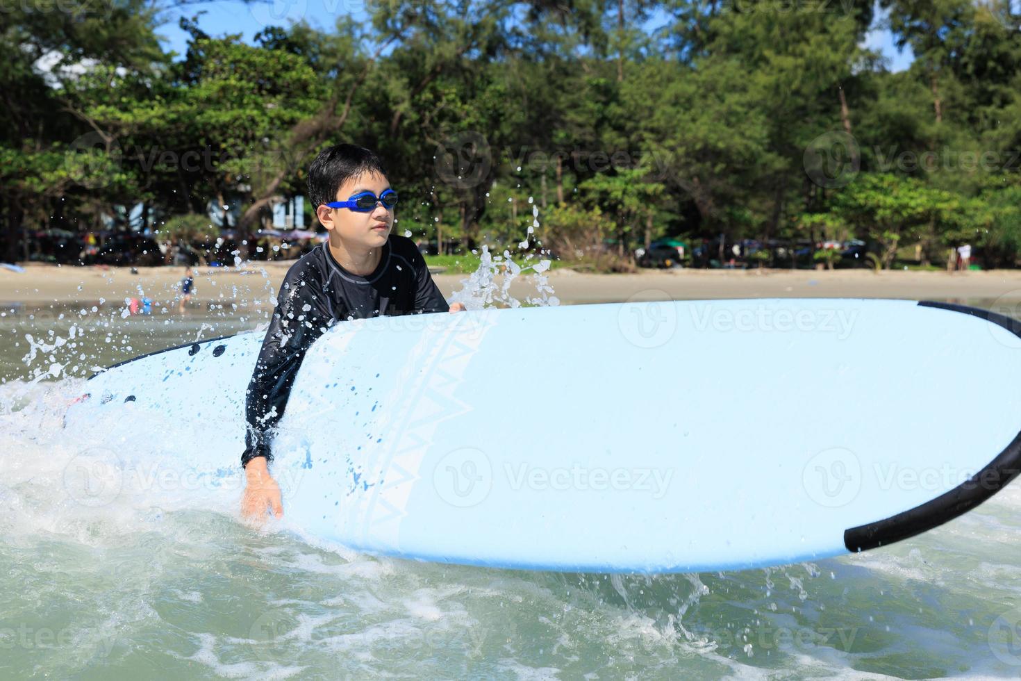 Young boy, student in surfing, holding softboard and trying to bring it back wading into sea to practice while playing against waves and splashing water. photo