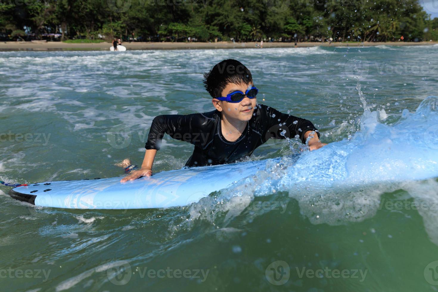 Young boy, student in surfing, holding onto softboard and trying to bring it back into sea to practice while playing against waves and splashing water. photo