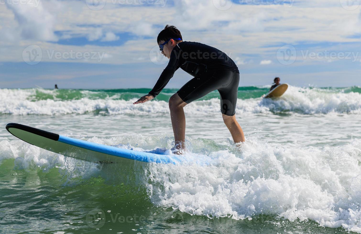 Young boy wearing swimming goggles stands on soft board while practicing surfing in beginner's class. photo