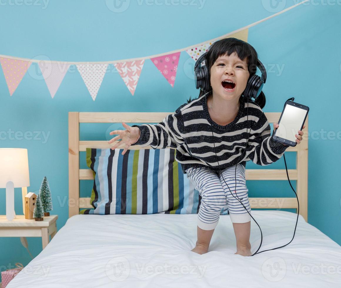 Cute little girl sing a song with smartphone in her bedroom, Happy asian child little girl listening the music with headphone on the bed, technology concept photo