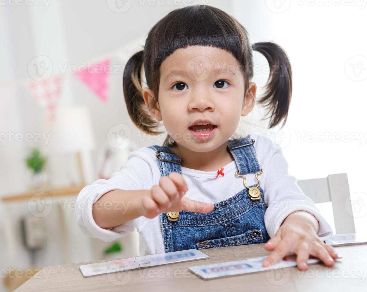 Little asian girl sitting at table in room, Preschooler girl playing the jigsaw on sunny day, kindergarten or day care. photo