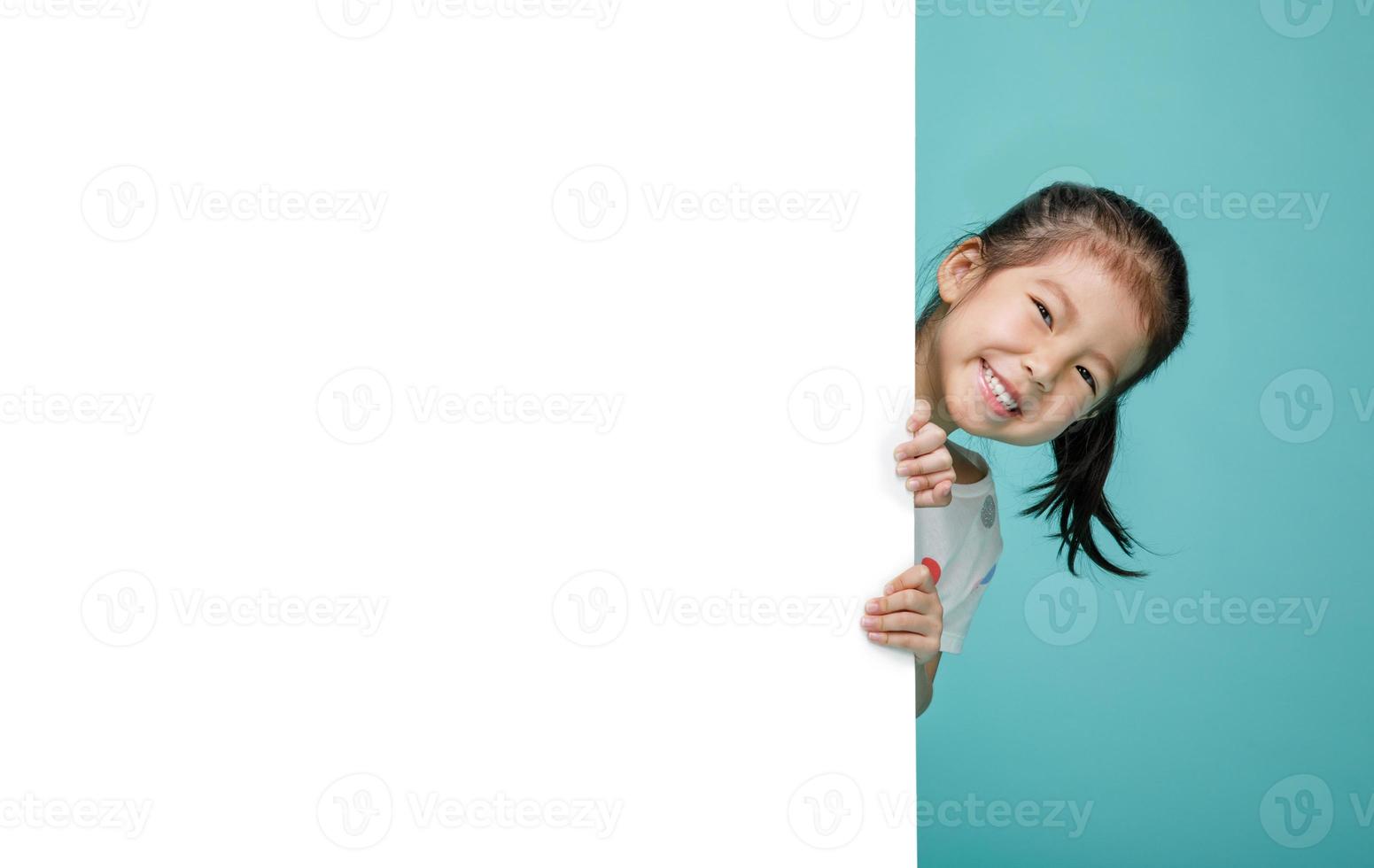 Smiling happy cute child hiding behind a blank white board, empty space in studio shot isolated on colorful blue background photo