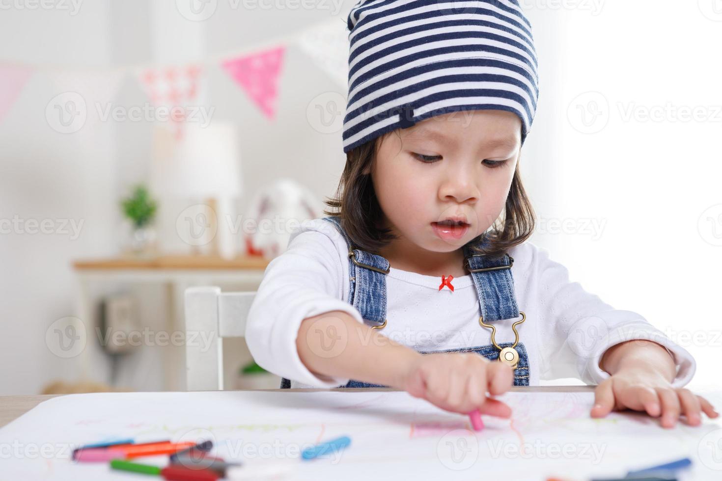 niña asiática sentada a la mesa en la habitación, niña preescolar dibujando en papel con bolígrafos coloridos en un día soleado, jardín de infantes o foto