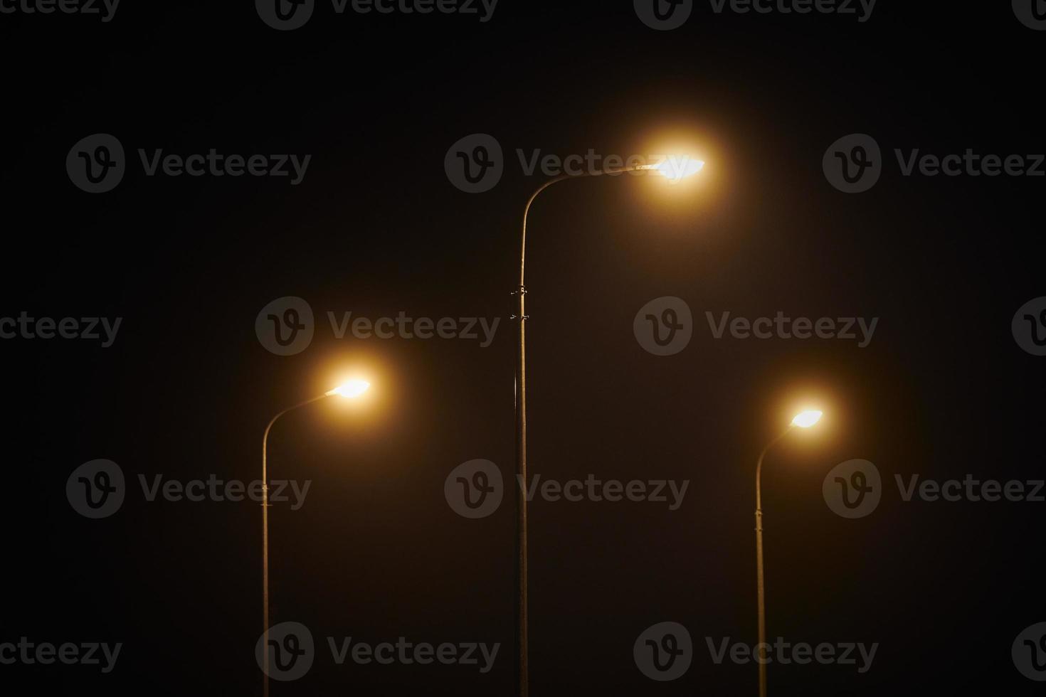 Three night lamppost shines with faint mysterious yellow light through evening fog at quiet night photo