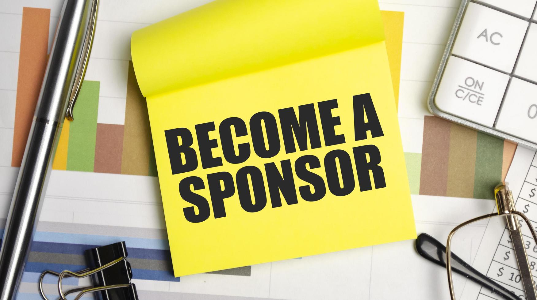 BECOME A SPONSOR text on the yellow paper with pen and glasses photo