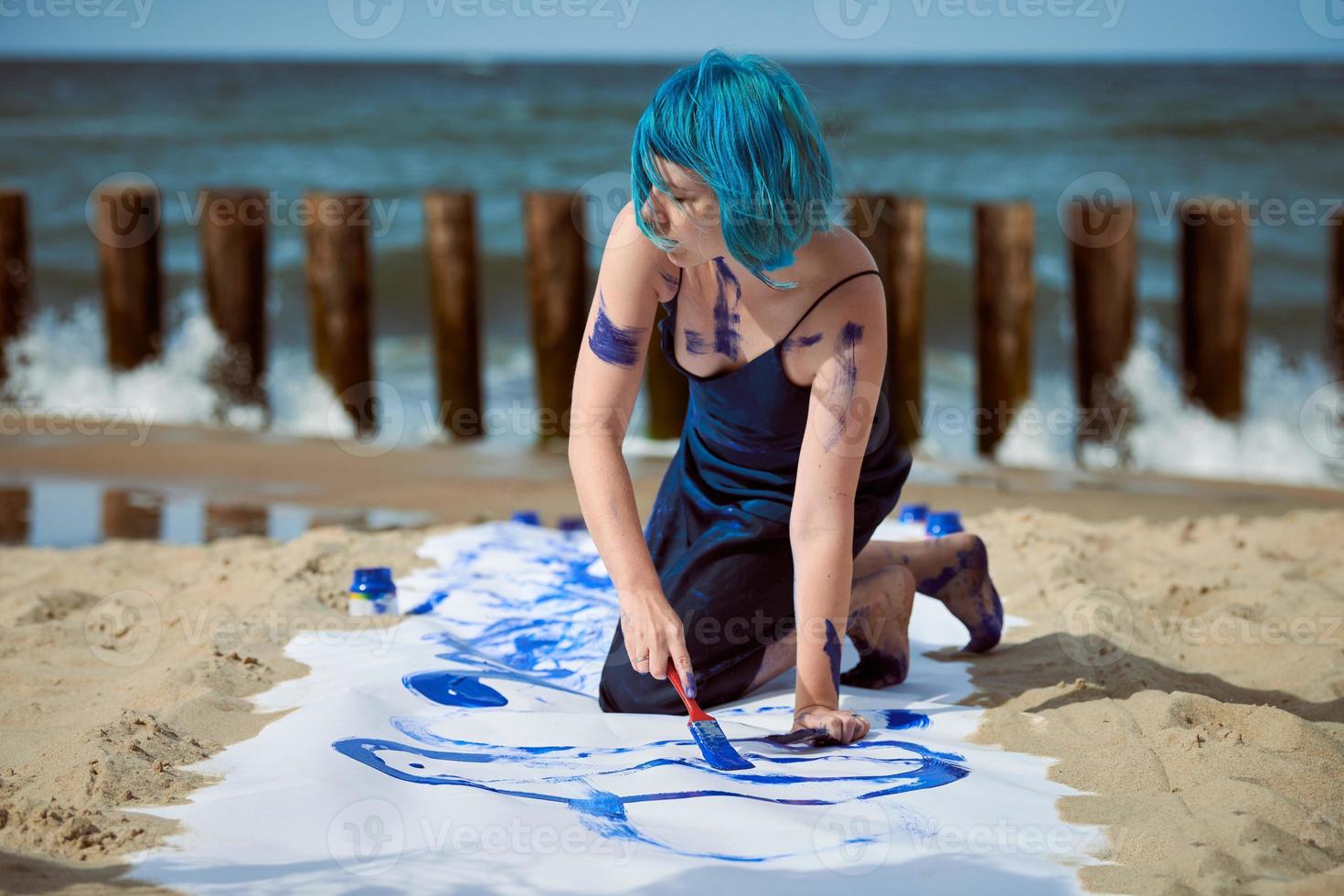 artista artística de cabello azul manchada con pinturas de gouache en un lienzo grande en la playa foto