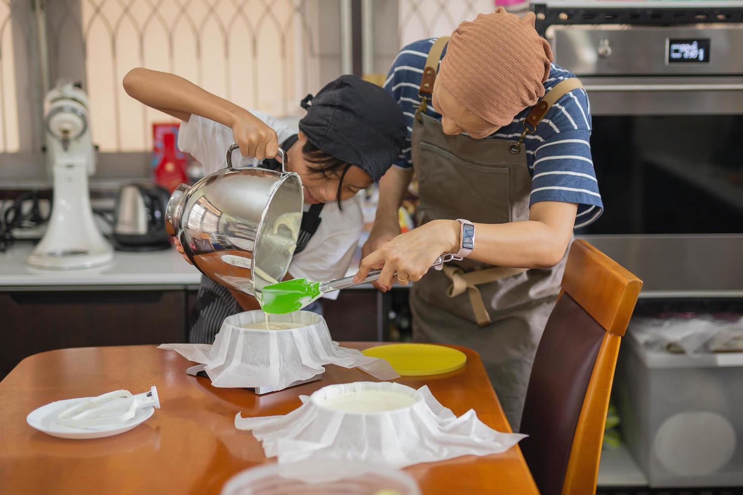 Mother with young son wearing apron pouring cream from mixing bowl into baking mold. photo