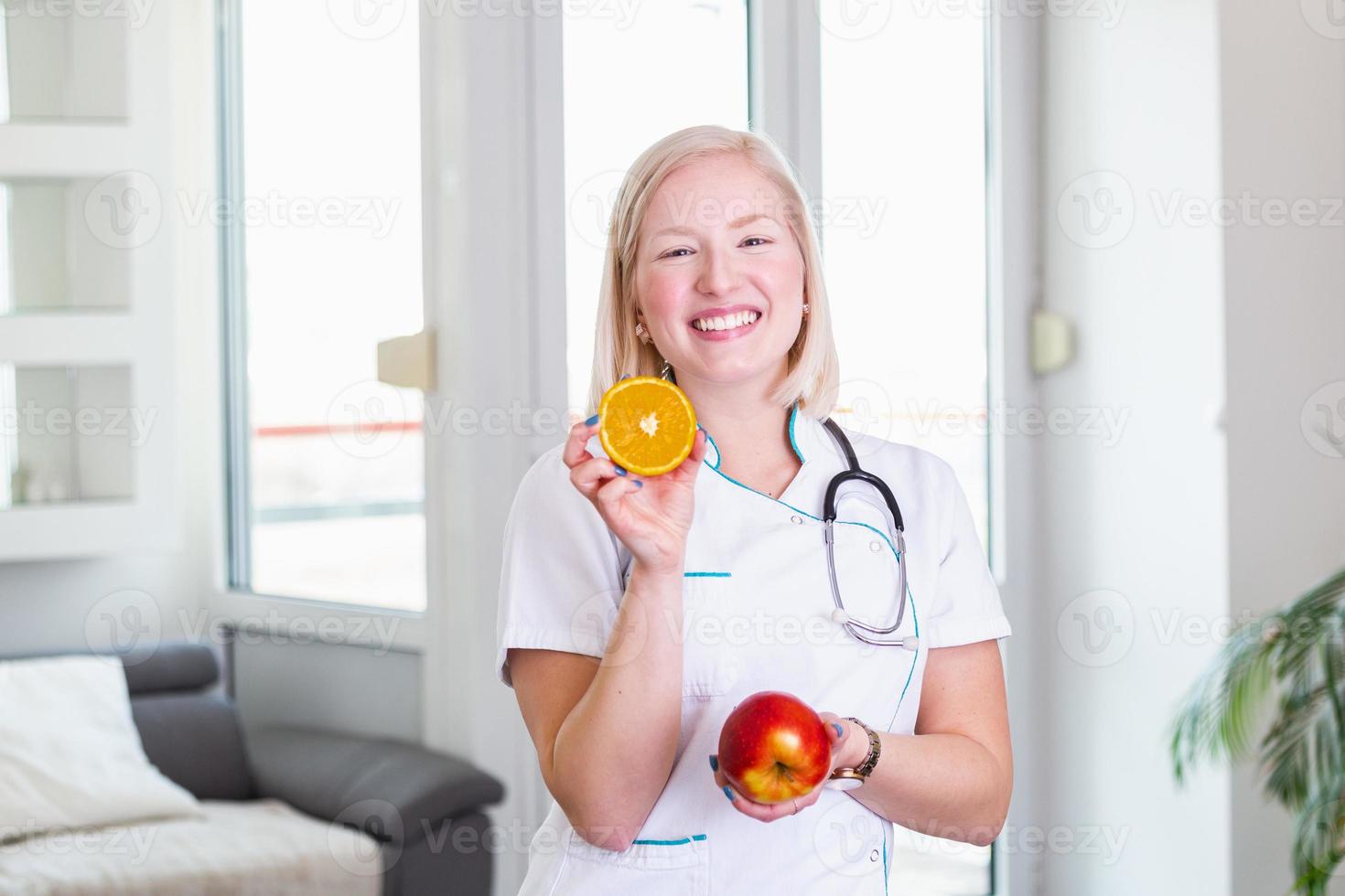 Smiling nutritionist in her office, she is holding a fruit and showing healthy vegetables and fruits, healthcare and diet concept. Female nutritionist with fruits photo