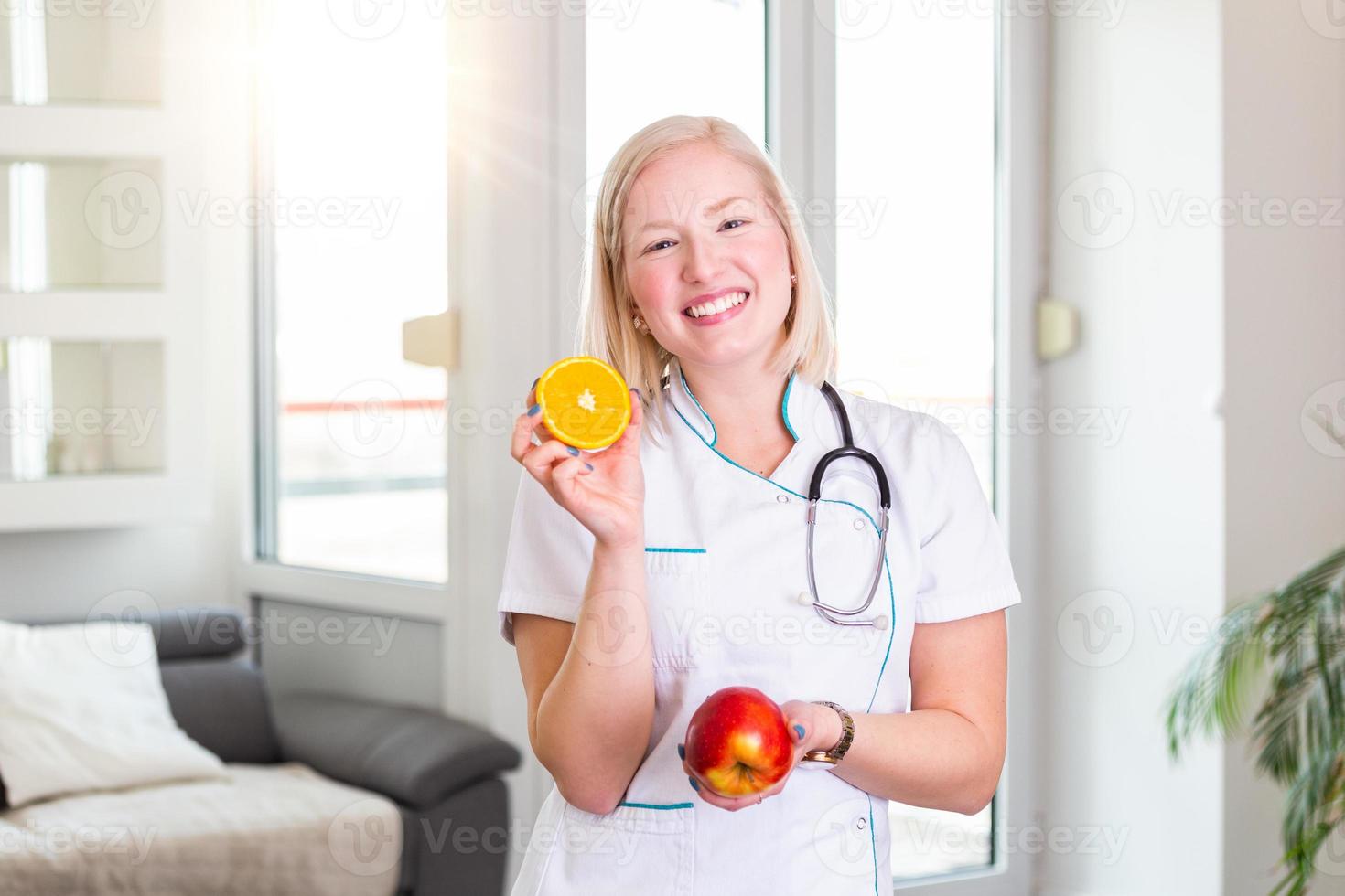 Smiling nutritionist in her office, she is holding a fruit and showing healthy vegetables and fruits, healthcare and diet concept. Female nutritionist with fruits photo