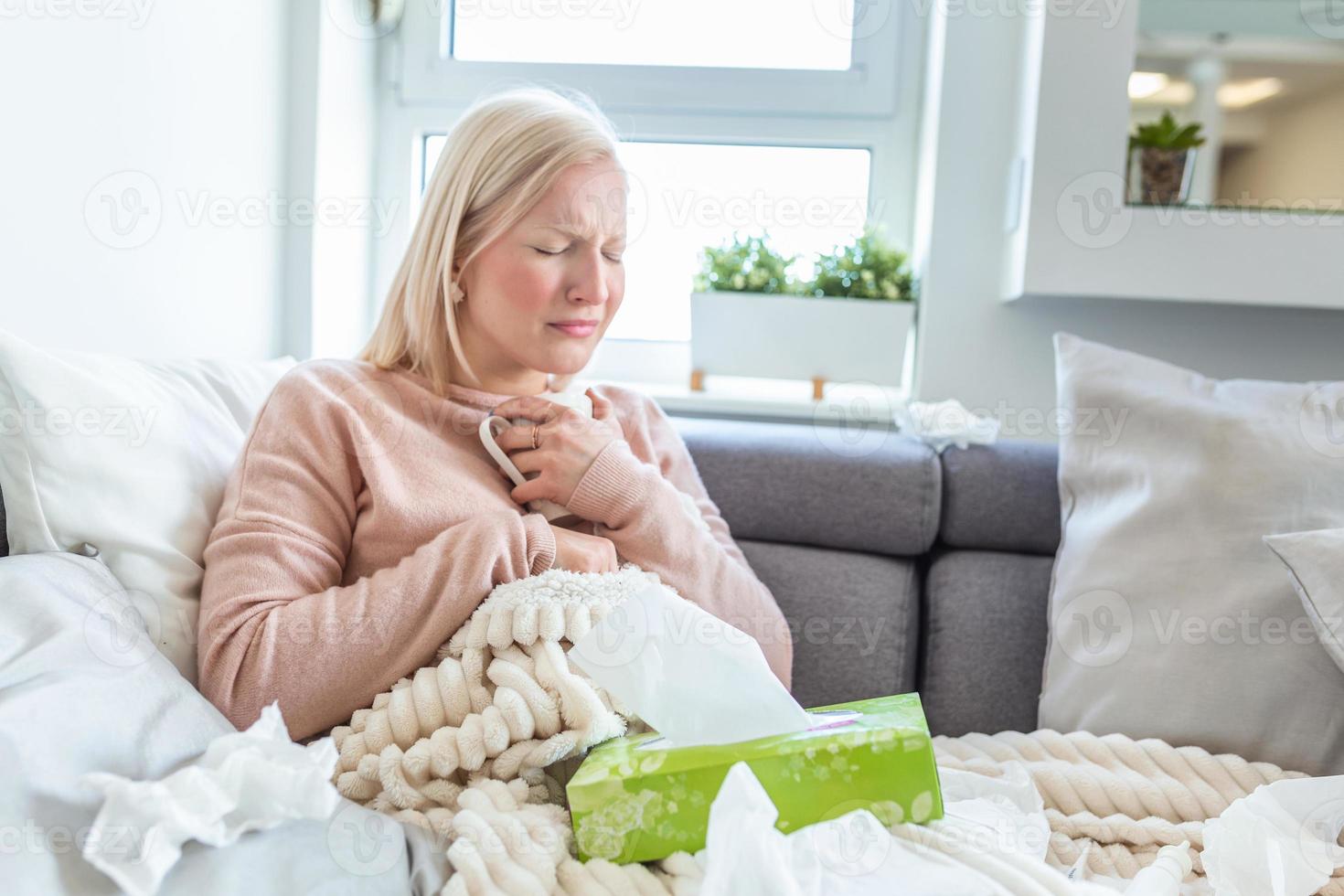 Young woman touching painful neck, sore throat for flu, cold and infection. Unhealthy Girl Covered In Blanket Wiping Nose. Healthcare Concept. Albino girl sick and drinking hot beverage photo