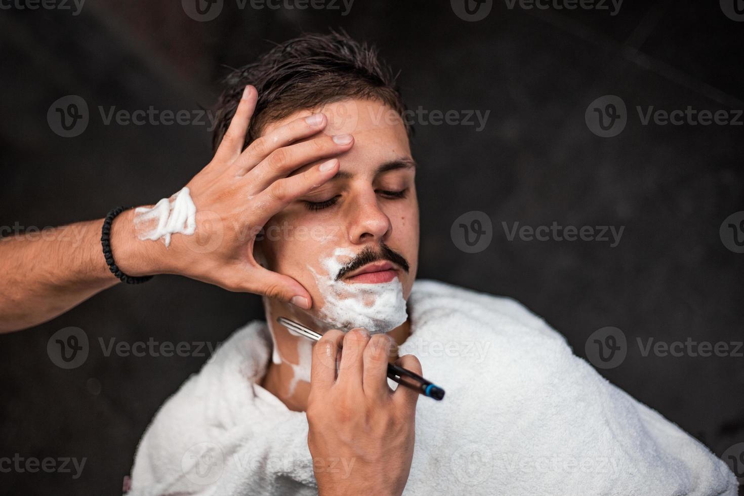 Man mith mustaches having a shave. Razor in hands of specialist barber. Young man getting an old-fashioned shave at the barber shop. Closeup, retro styled imagery photo