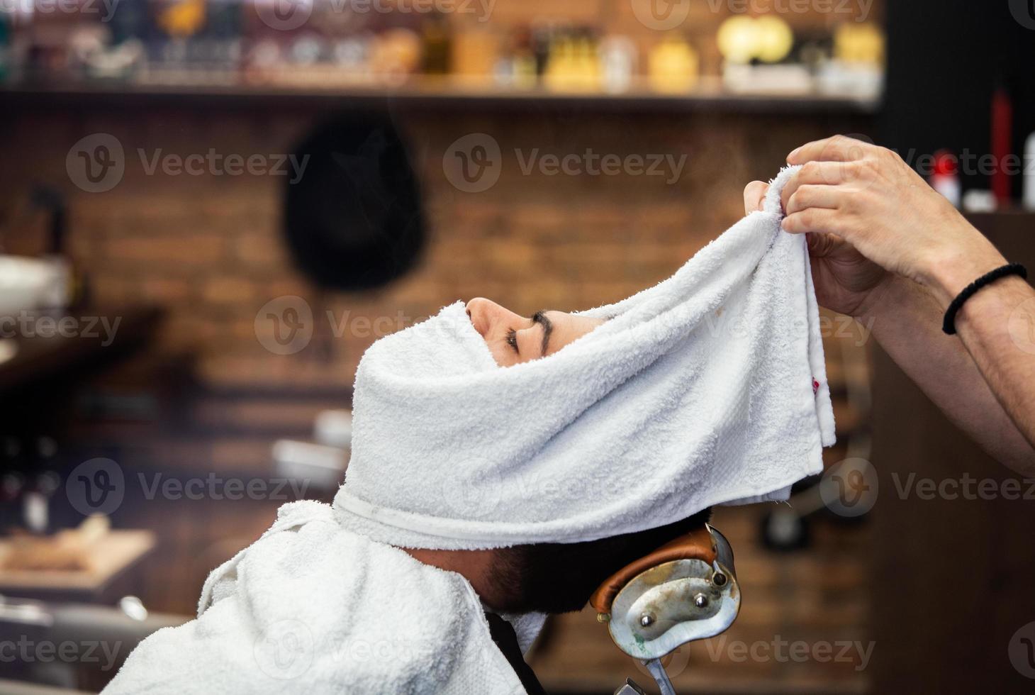Barber covers the face of a man with a hot towel. Traditional ritual of shaving the beard with hot and cold compresses in a old style barber shop. photo