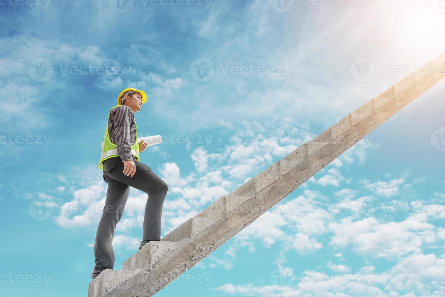 El ingeniero de construcción de un hombre de negocios asiático sostiene un plano de papel subiendo una escalera con un cielo azul, un crecimiento profesional y un concepto de éxito foto