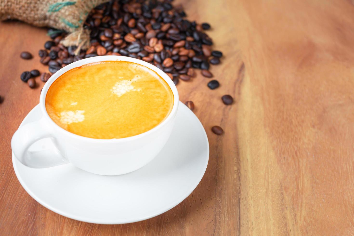 Close-up of a cup of latte with golden foam and mixed or blend coffee beans in a brown sack on an old wooden floor, top view. photo