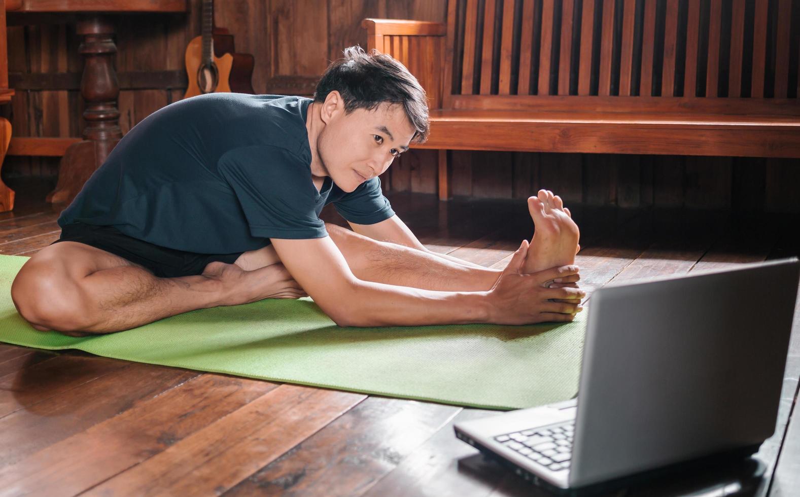 young Asian man  coach internet video online training yoga instructor modern laptop screen meditate Sukhasana posture relax breathe easy seat pose gym healthy lifestyle at home concept. photo