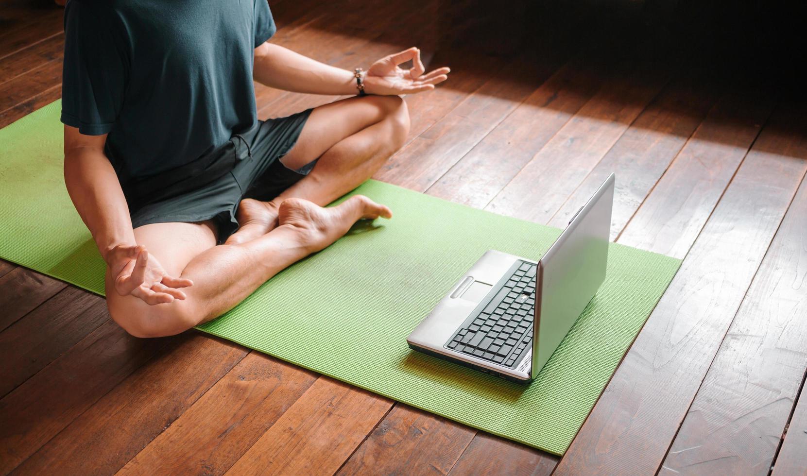 young Asian man  coach internet video online training yoga instructor modern laptop screen meditate Sukhasana posture relax breathe easy seat pose gym healthy lifestyle at home concept. photo