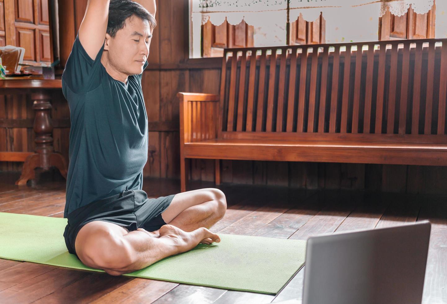 young Asian man  coach internet video online training yoga instructor modern laptop screen meditate Sukhasana posture relax breathe easy seat pose gym healthy lifestyle at home concept. photo