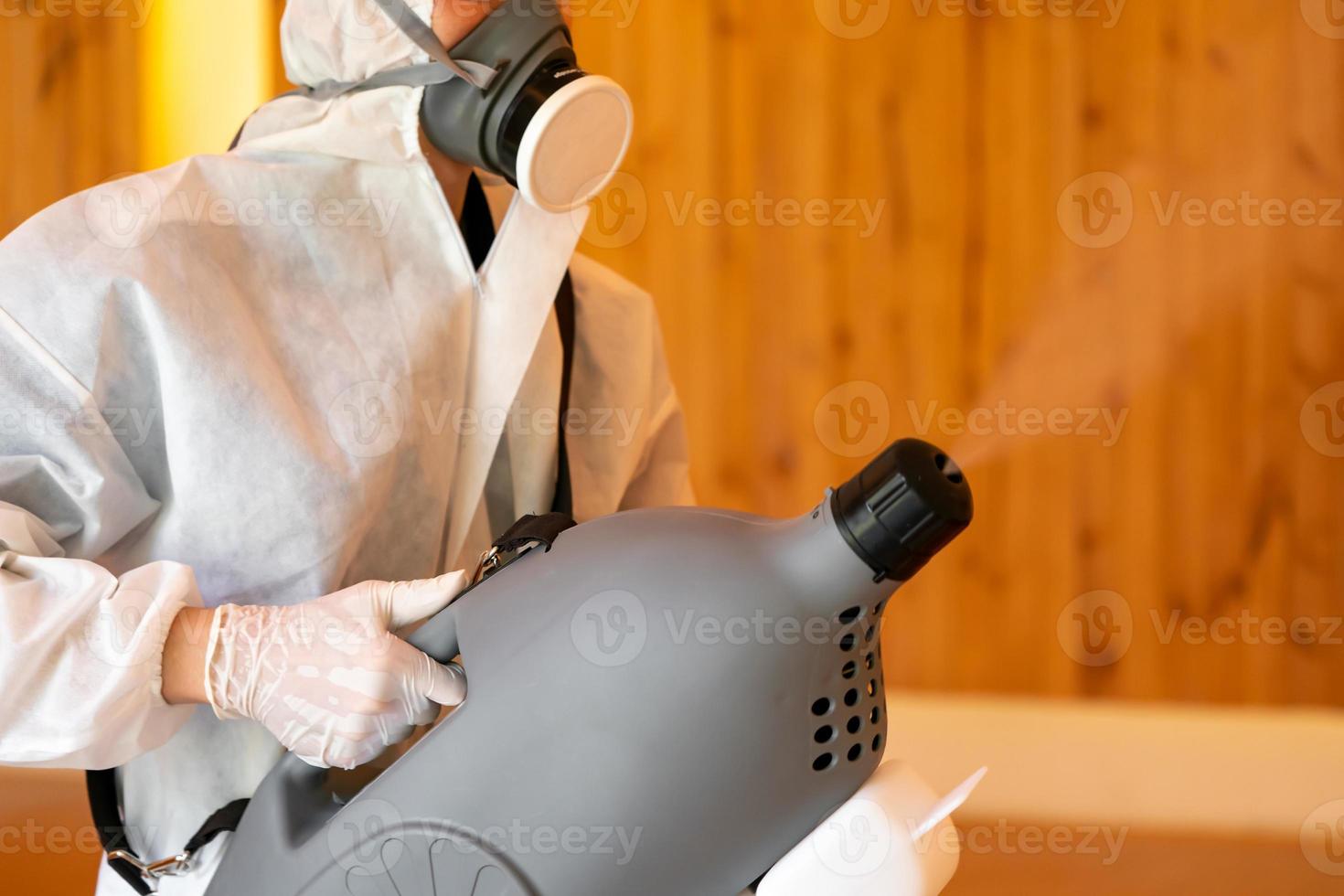 Professional technical man in prevention suit sprays sterilising solution by electrical spray machine on the wood floor and white background with studio light. photo