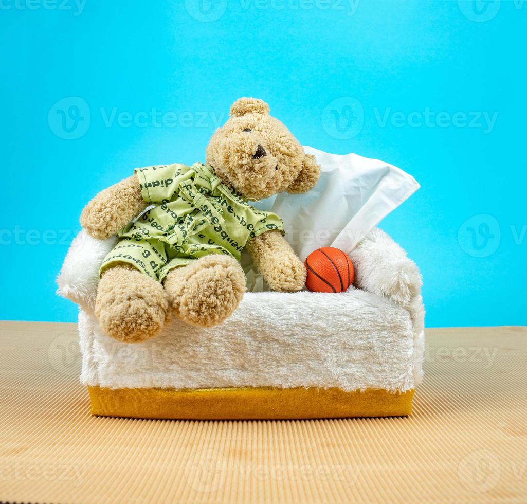 el oso de peluche se pone el pijama y se sienta en la caja de pañuelos con el baloncesto al lado foto