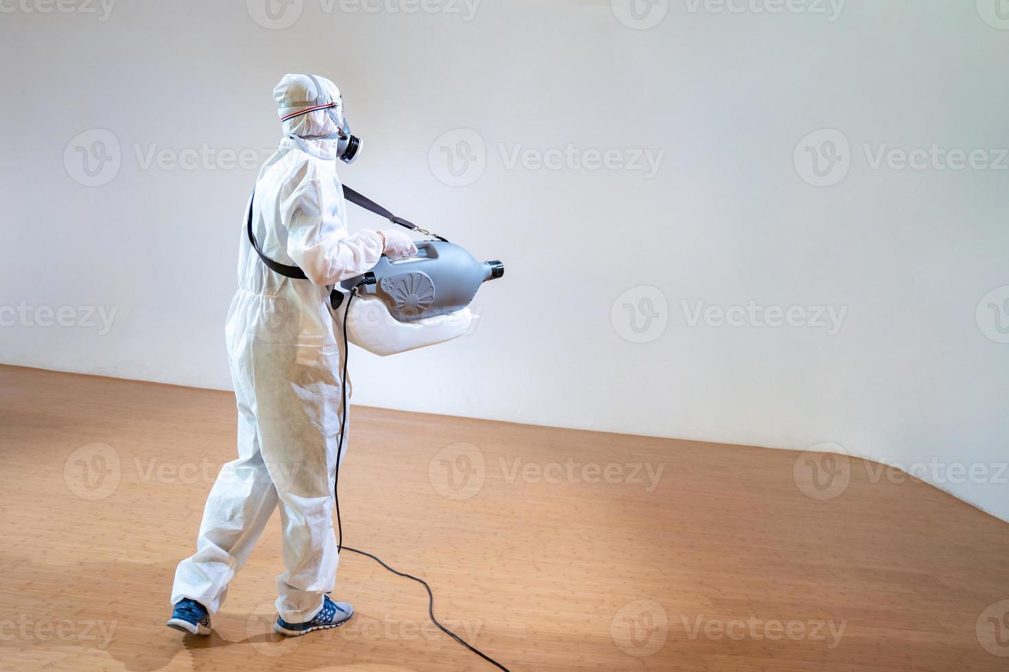 Professional technical man in prevention suit sprays sterilising solution by electrical spray machine on wood floor and white background with studio light. photo