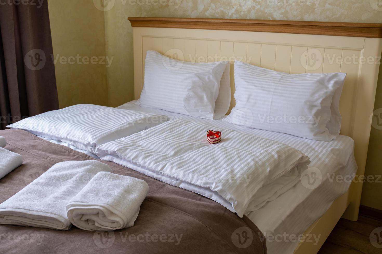 A small gift box in the shape of a heart on a white bed in a hotel room. photo