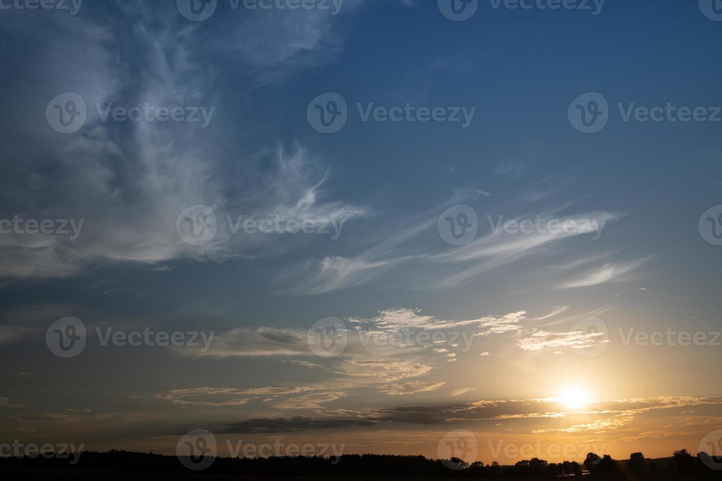 The sun goes down on the horizon. The sky is blue with white clouds. The sky glows red on the horizon. The outlines of trees can be seen at the bottom. photo
