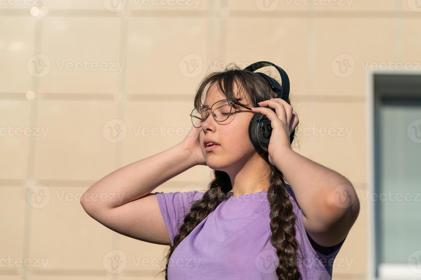 a teenager girl stands on the street, presses headphones to her ears with her hands and listens to music with her eyes closed. photo