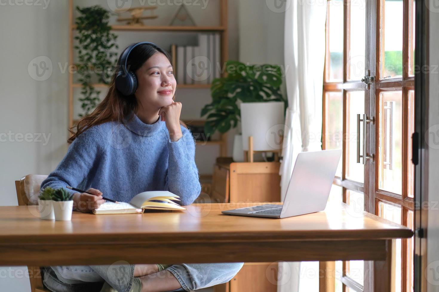 Smiling girl in headphones sitting at a desk looking outside and using a laptop to study online. Smart young women who are happy in headphones take courses on the web or practice using computers. photo