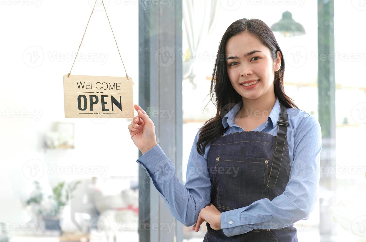 una mujer de negocios feliz asiática es una camarera en un delantal, el dueño del café se para en la puerta con un cartel abierto esperando a los clientes. concepto de pequeña empresa, cafés y restaurantes foto