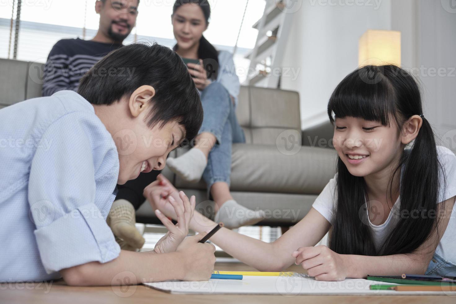 Asian Thai siblings are lying on living room floor, drawing homework with colored pencils together, parents leisurely relax on a sofa, lovely happy weekend activity, and domestic wellbeing lifestyle. photo