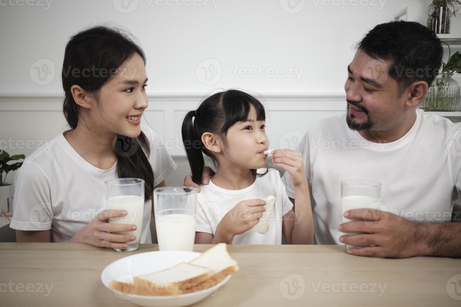 A Healthy Asian Thai family, a little daughter, and young parents drink fresh white milk in glass and bread joy together at a dining table in morning, wellness nutrition home breakfast meal lifestyle. photo