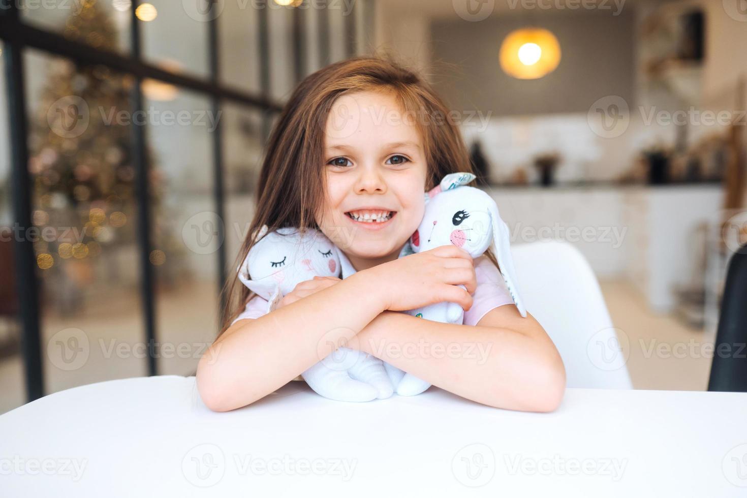 Little smiling girl hugging favorite toys while sitting at table photo