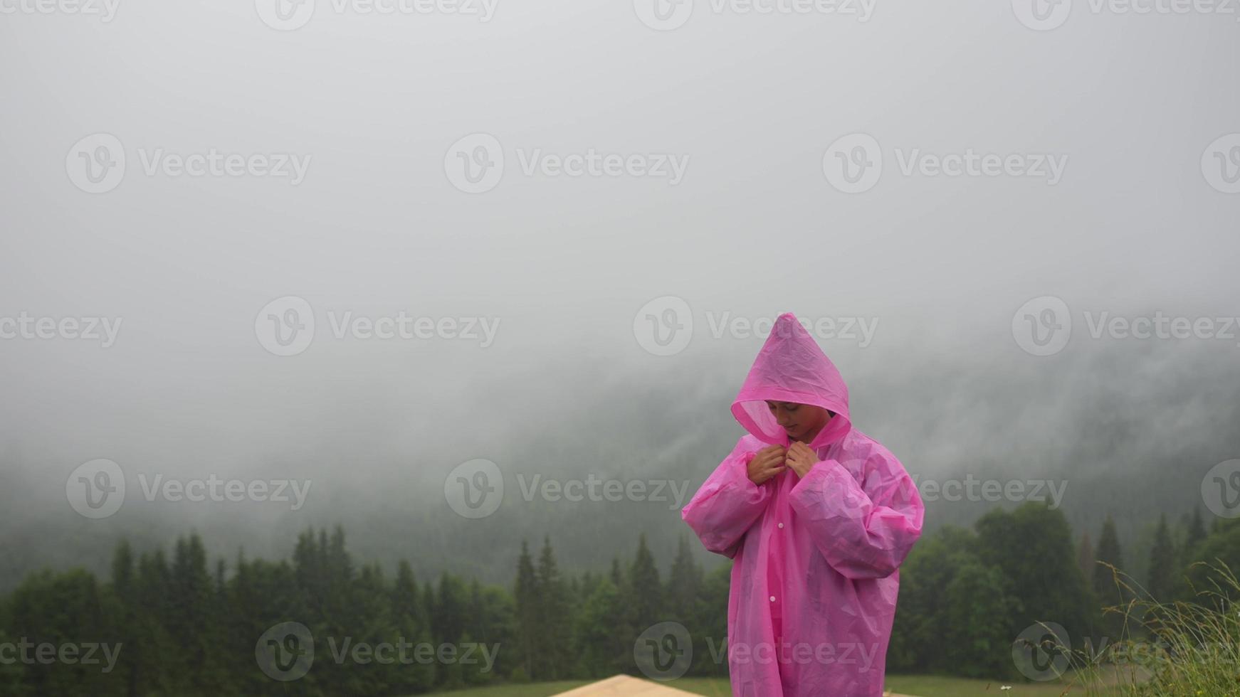 Attractive hiker girl in a pink raincoat stands on a mountain photo
