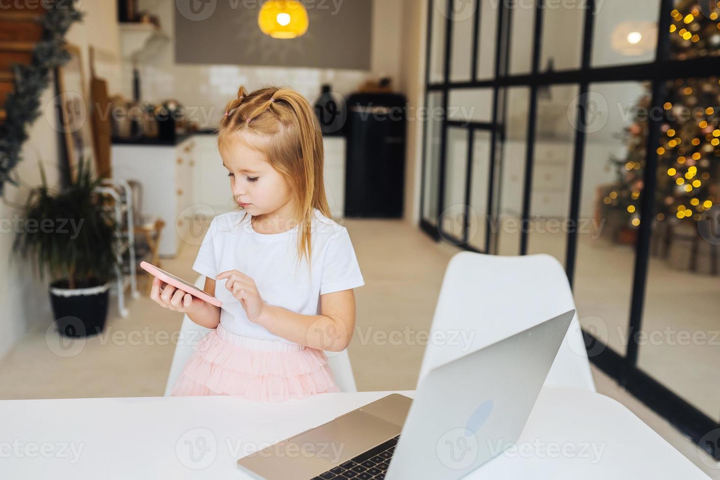 Little kid sit at desk with laptop hold mobile phone photo