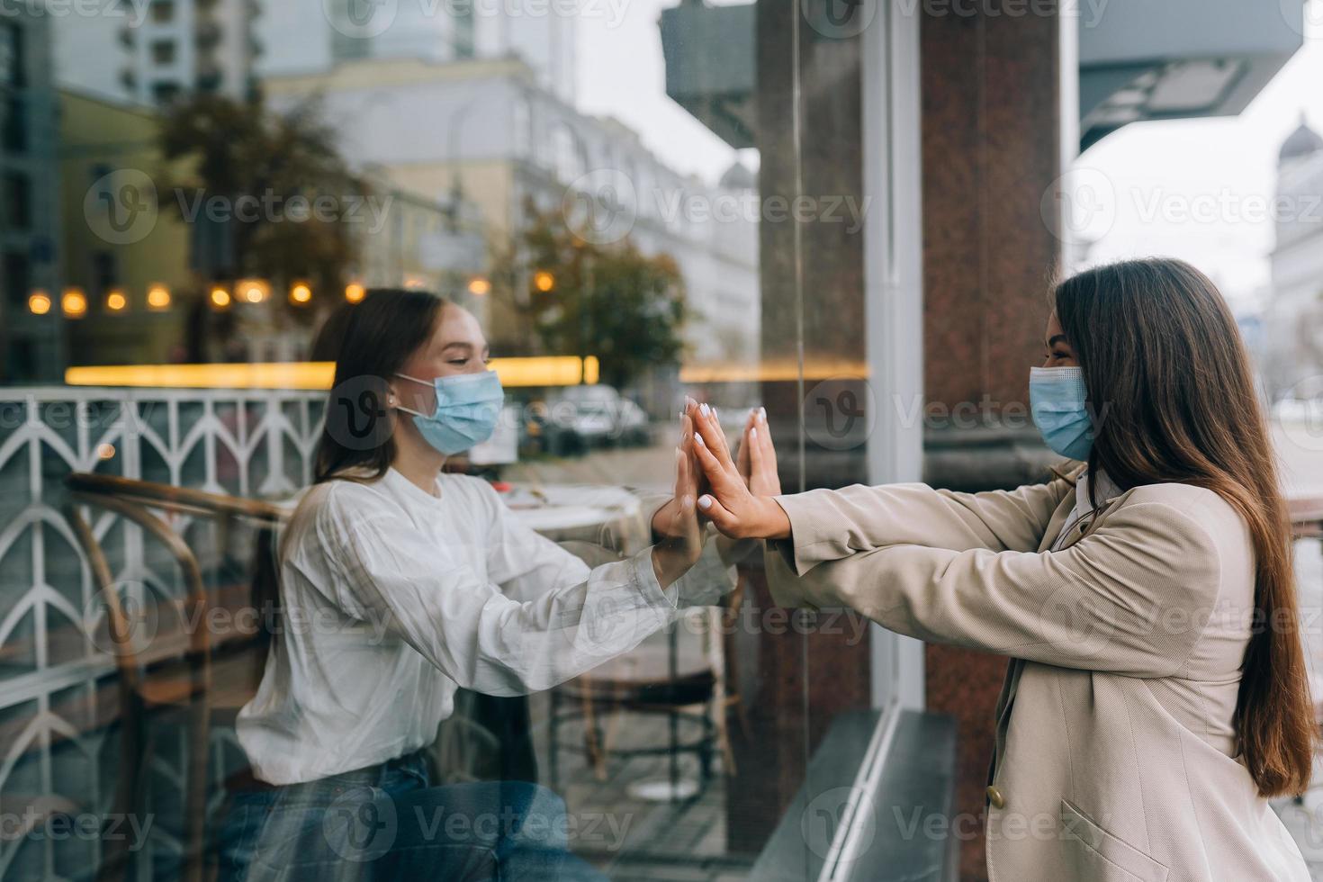 Two women in protective masks opposite each other, window between them. photo