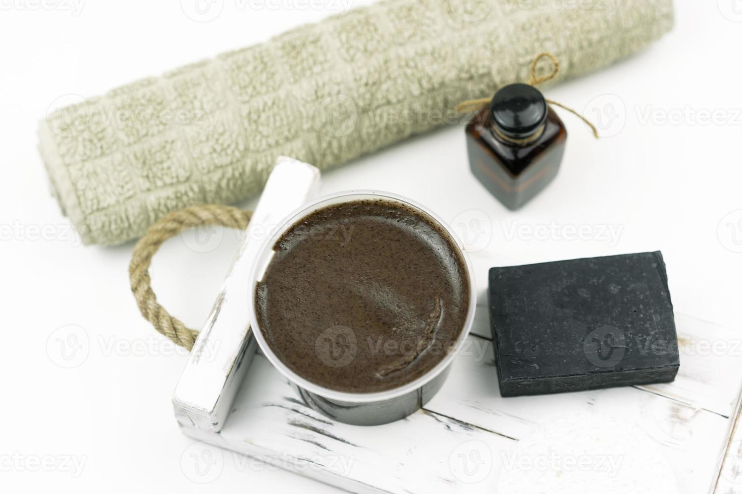 A chocolate scrub for body, a black hand made soap, a cooton towel, a natural oil for face stand on the white wooden tray in a modern bathroom photo