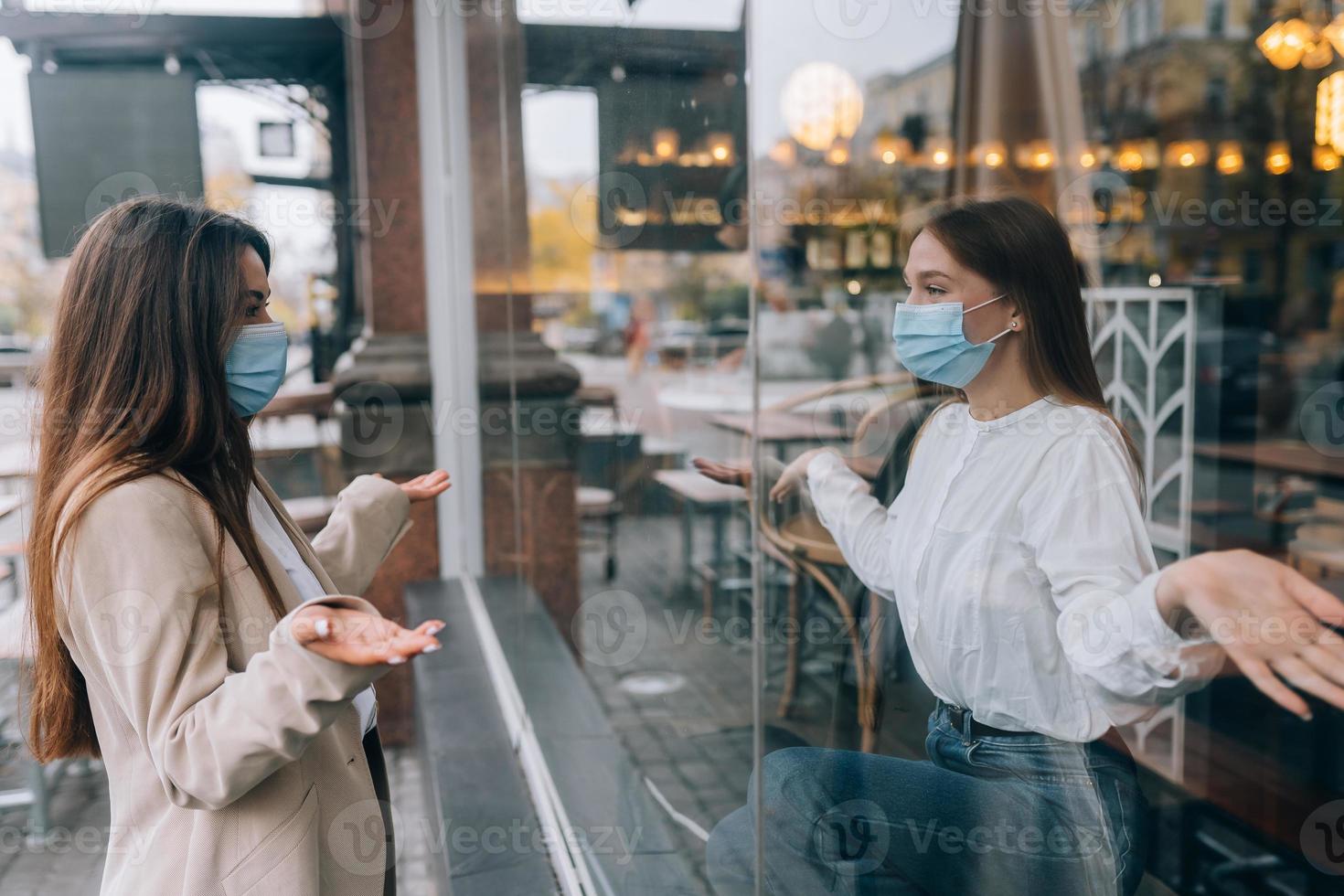Two women in protective masks opposite each other, window between them. photo