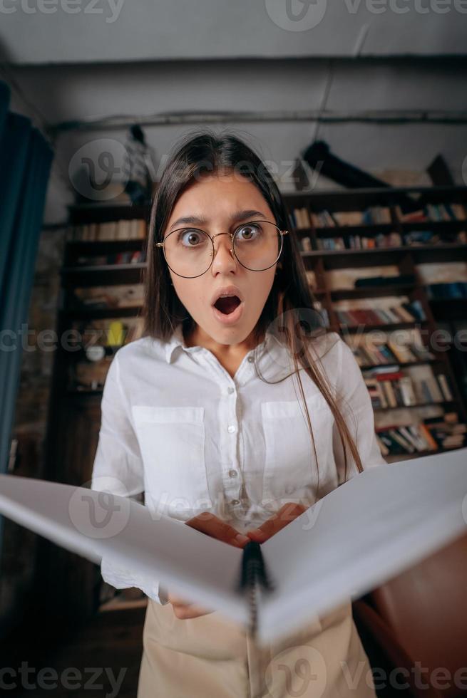 Young beautiful woman in eyeglasses looks shocked after reading a book. Looking at camera. photo
