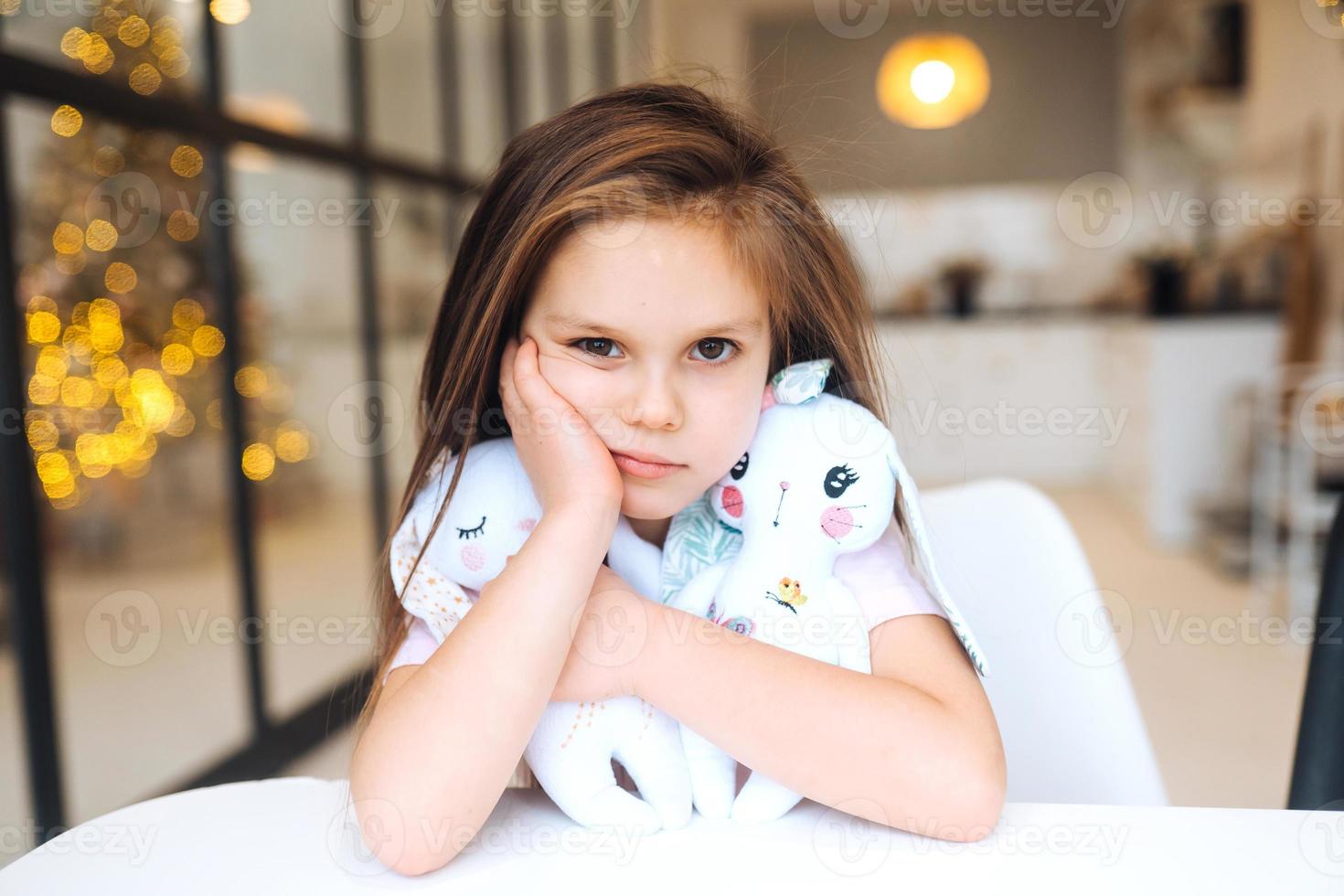 Little sad girl hugging favorite toy while sitting at table photo