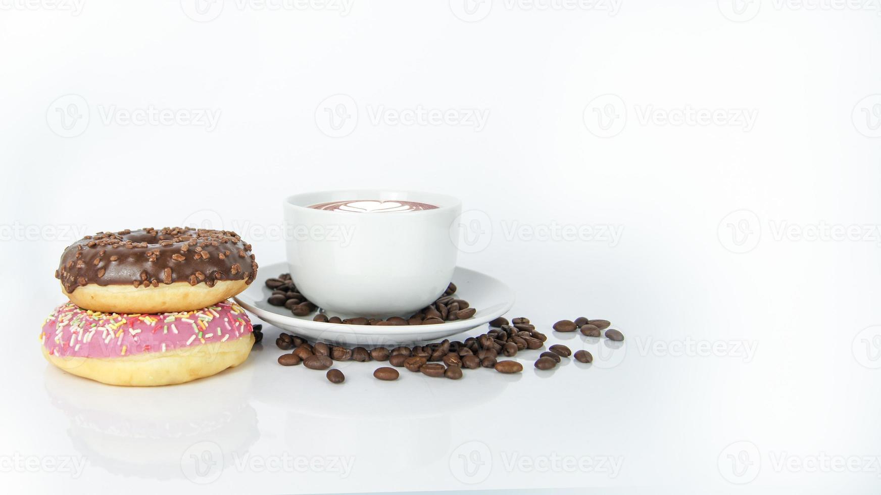 Aromatic coffee latte with leaf on the top in white cup on plate covered by brown seeds next to doughnuts. Chocolate and raspberry snacks on white background. Isolated objects in composition photo