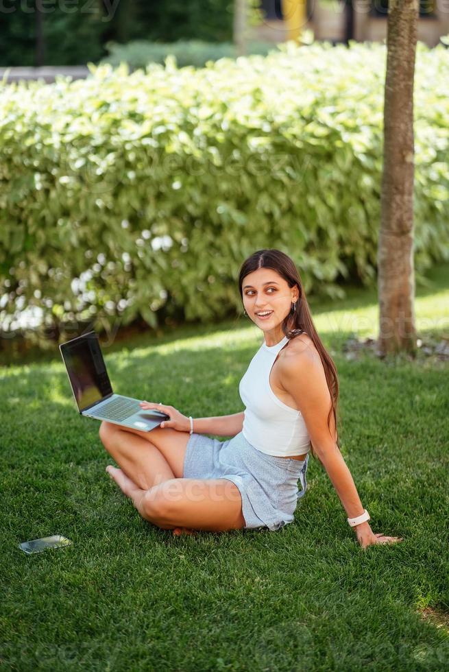 la joven trabaja con una laptop, sentada en el césped. foto