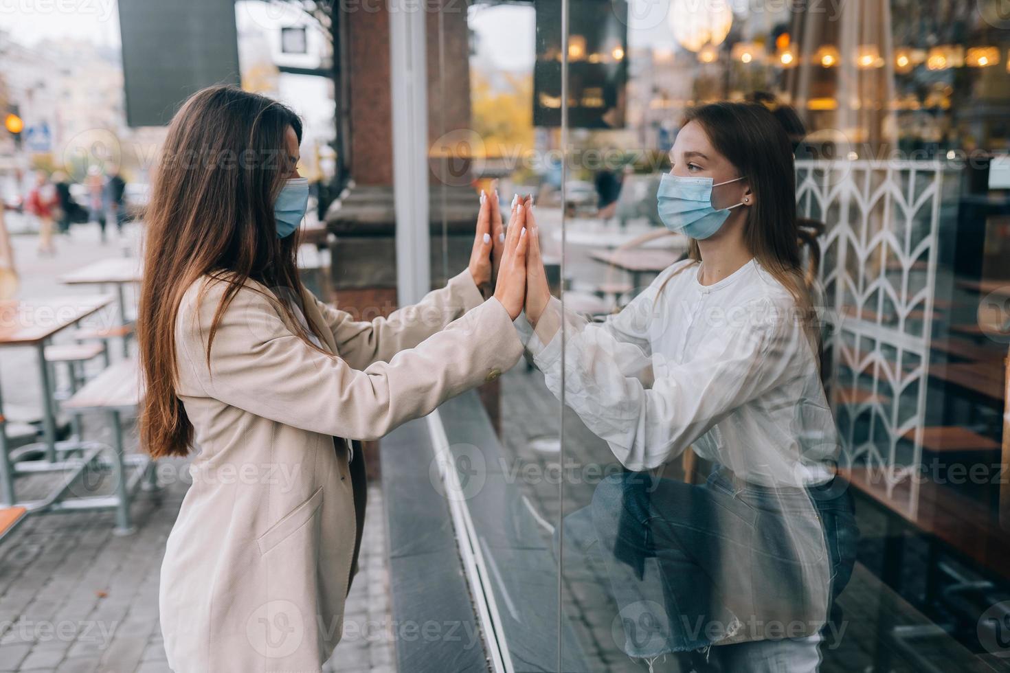Two women in protective masks opposite each other, window between them. photo