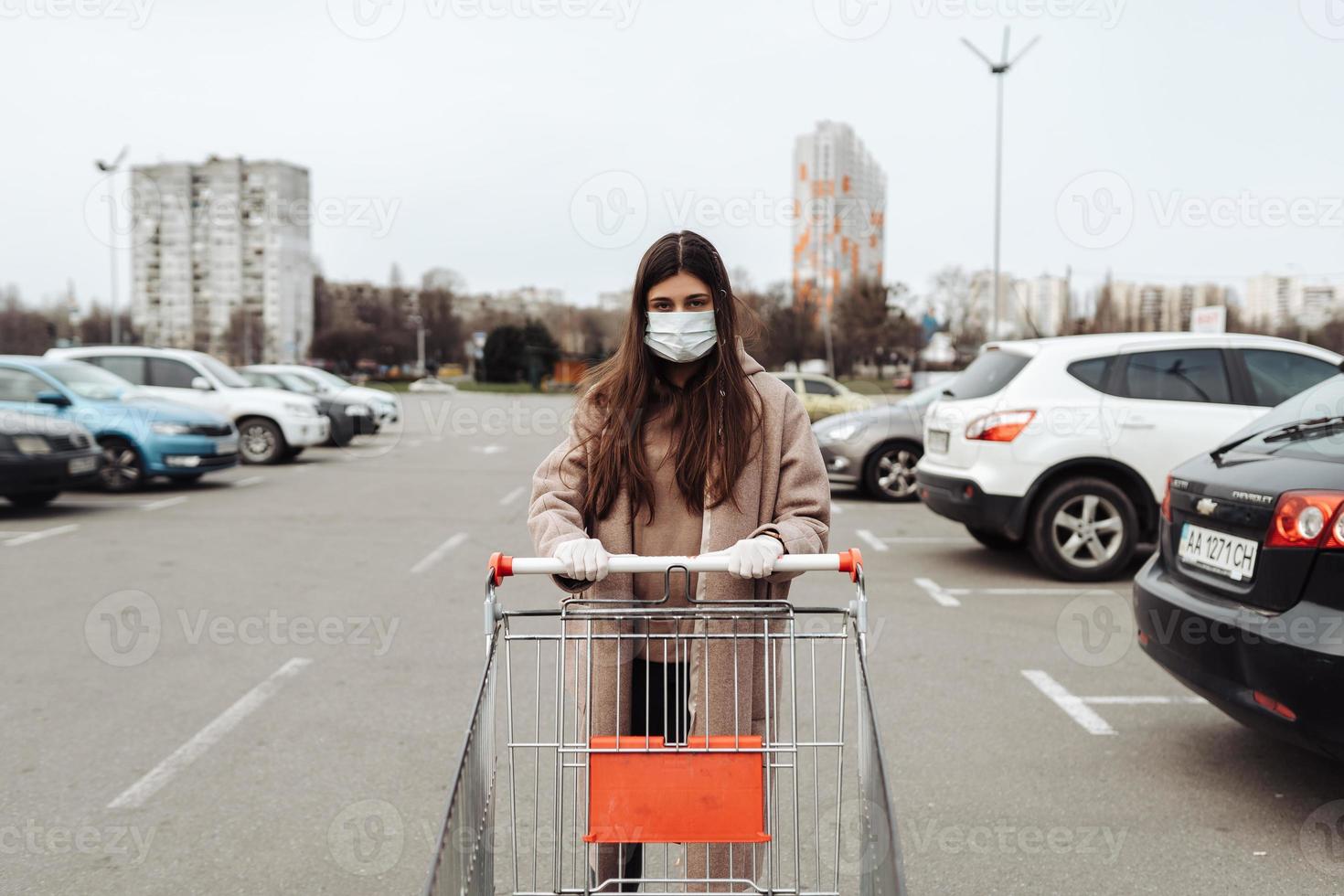 mujer joven con mascarilla de protección contra el coronavirus 2019-ncov empujando un carrito de compras. foto