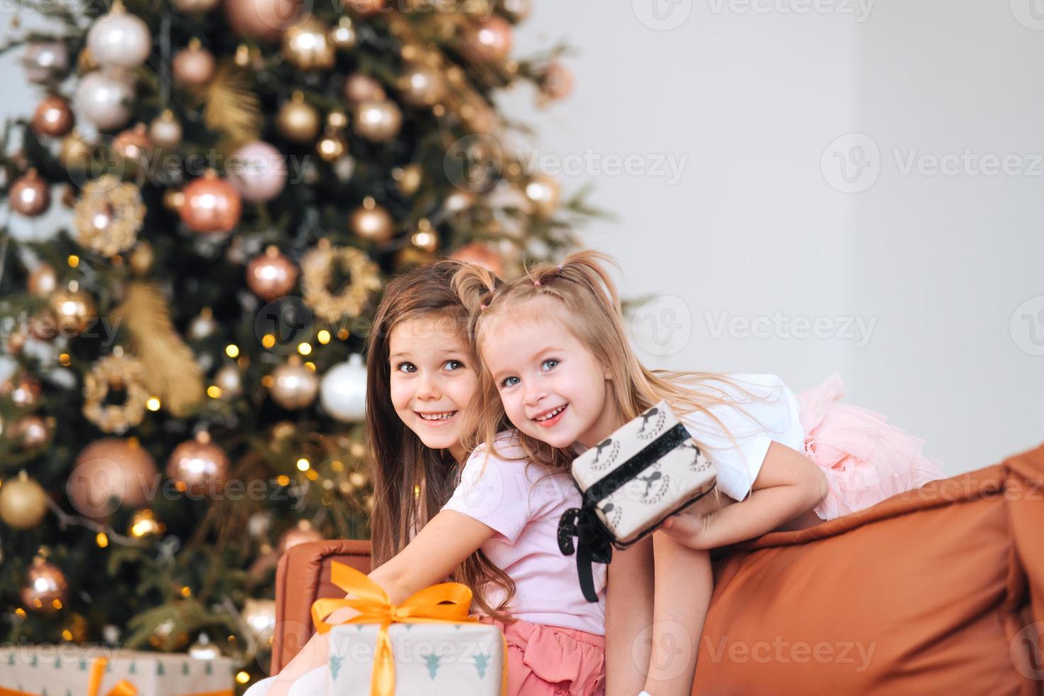 Two little girls are playing with New Year's gifts by the tree at sofa. photo