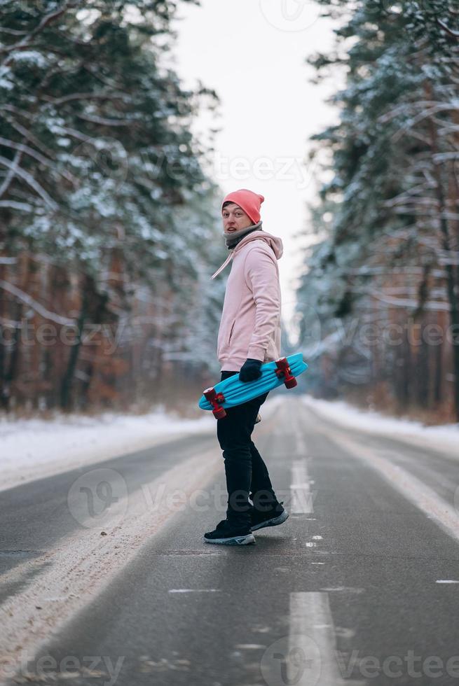 Skateboarder standing on the road in the middle of the forest, surrounded by snow photo