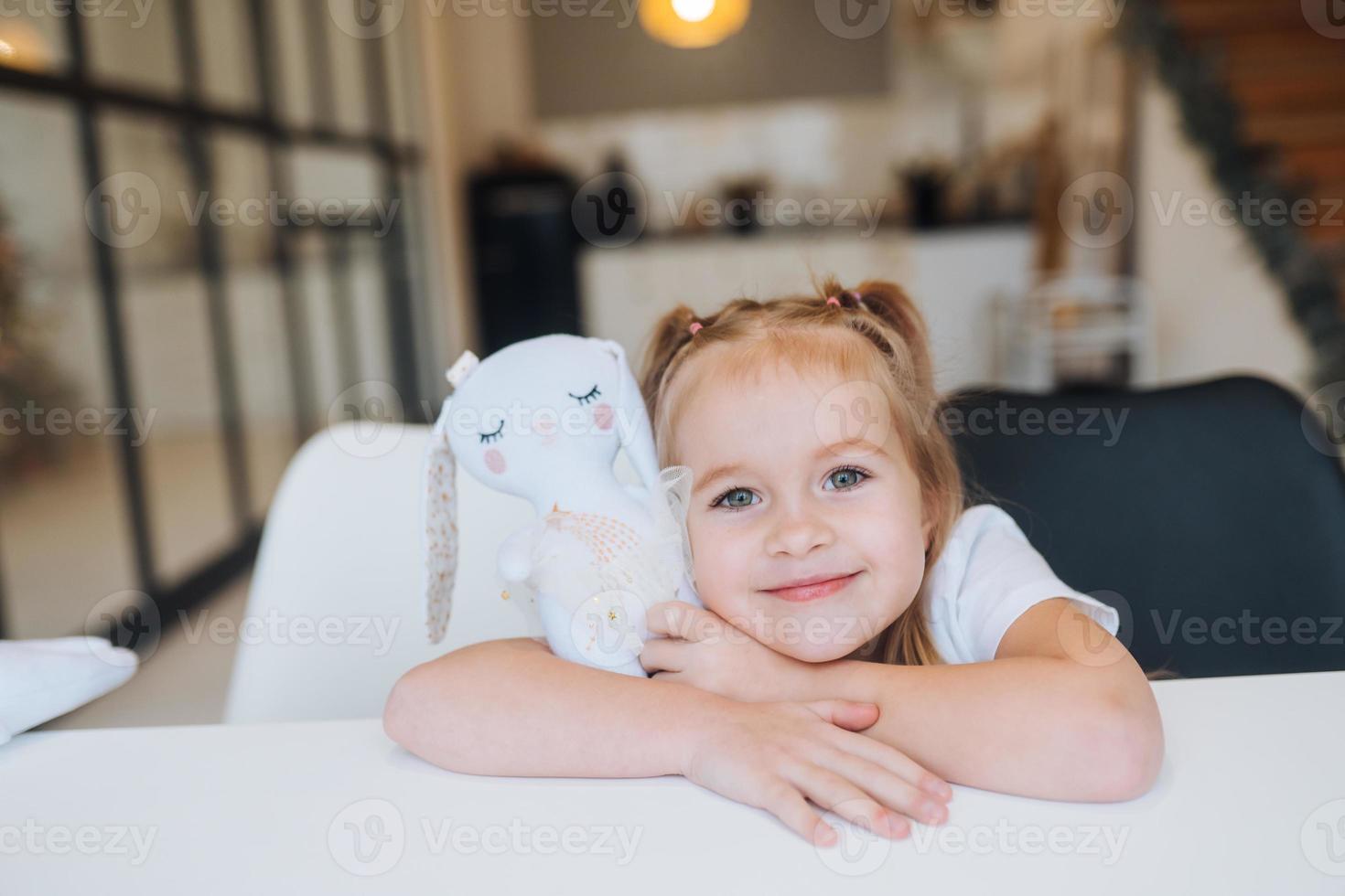 Little smiling girl hugging favorite toys while sitting at table photo