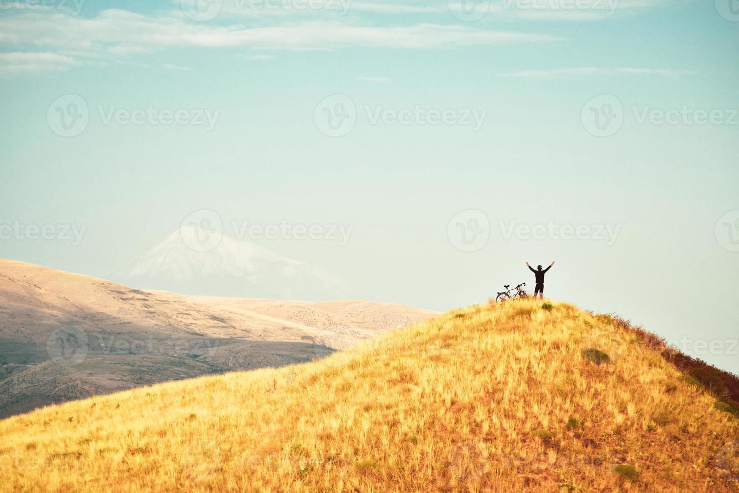 Top view inspirational excited joyful caucasian male cyclist stand on hilltop by red touring bicycle excited enjoy Ararat mountains background. Solo cycling adventure lifetime photo