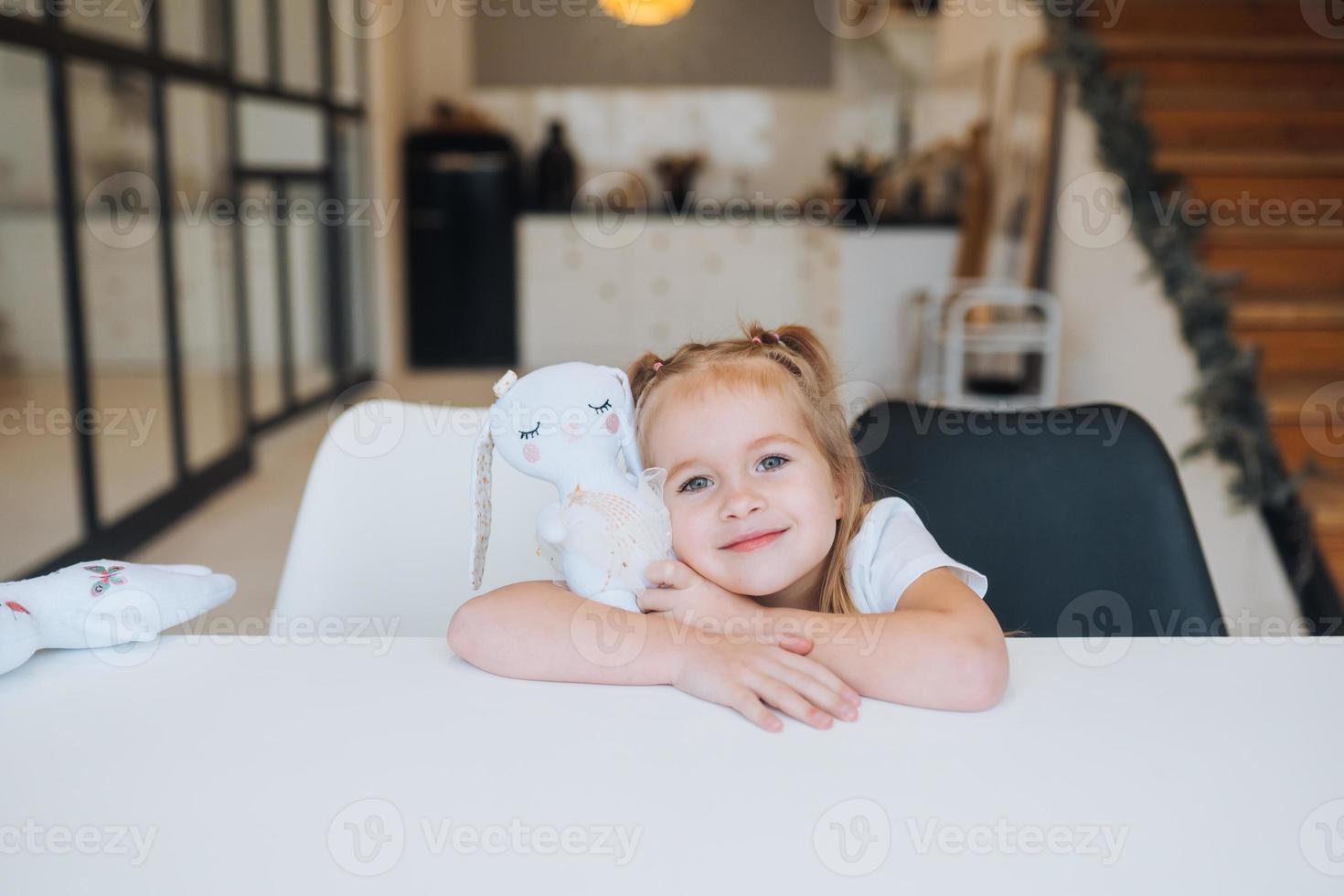 Little smiling girl hugging favorite toys while sitting at table photo