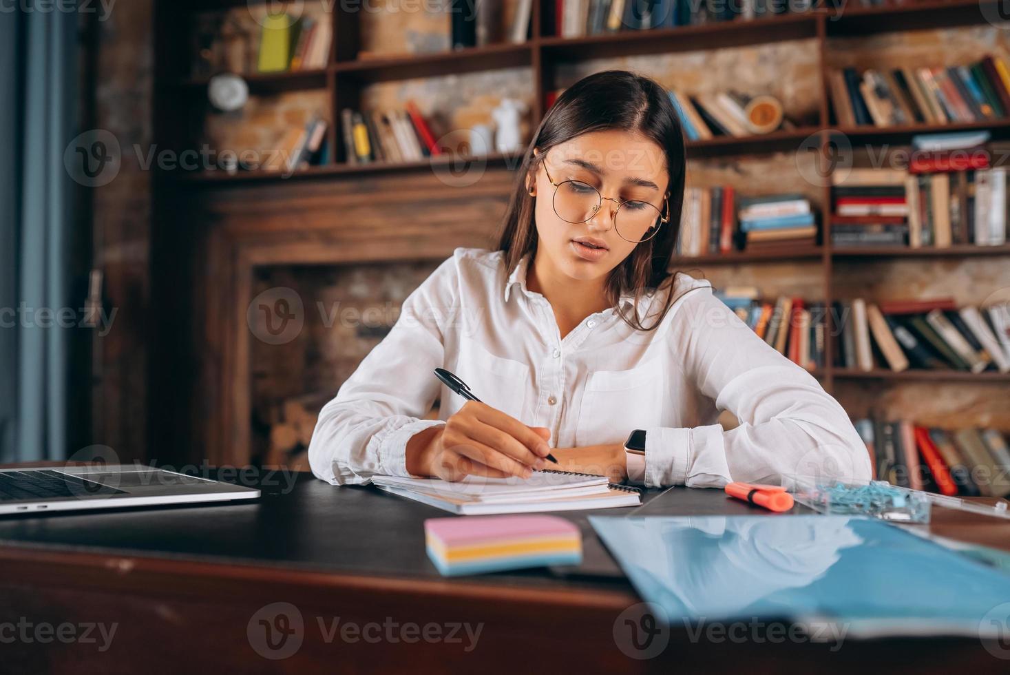 una joven con anteojos escribe en un cuaderno mientras se sienta en la mesa foto